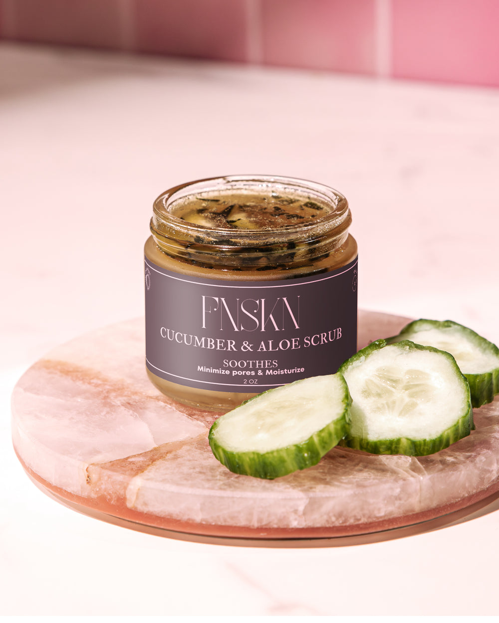 A jar of Cucumber & Aloe Scrub with sliced cucumbers and a whole aloe leaf on a pink salt block, placed on a pink surface.