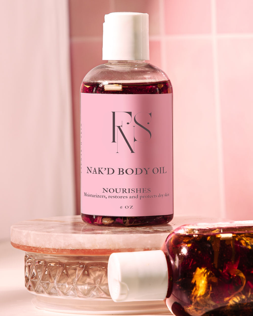 A bottle of NAK’D BODY OIL with pink packaging, placed on a marble surface, with a soft, warm background suggesting a bathroom setting.