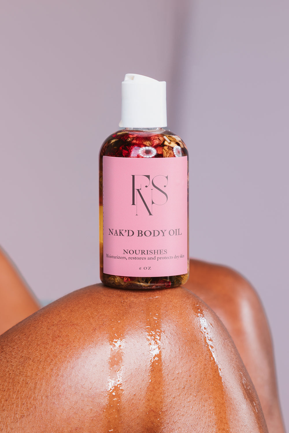 A bottle of NAK’D BODY OIL with pink packaging, placed on a marble surface, with a soft, warm background suggesting a bathroom setting.